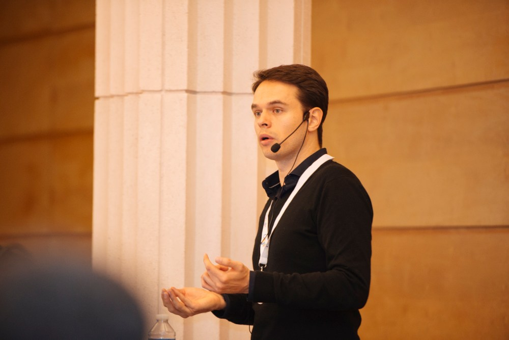 Un homme en train de donner une présentation avec un microphone sur l'oreille, gesticulant avec ses mains devant un podium.